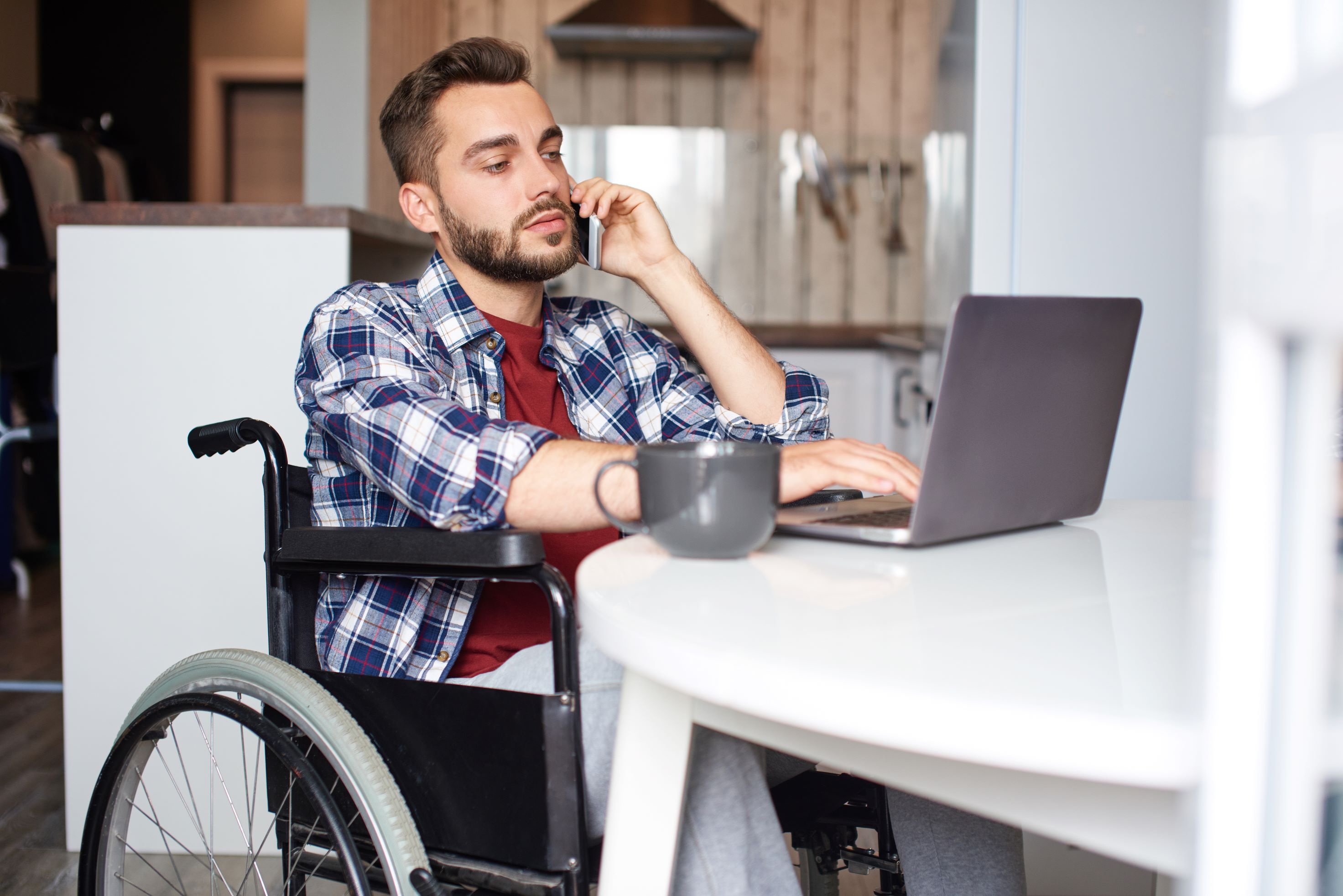 Young man who uses wheelchair sits at his laptop with smartphone in hand, waiting for customer service.